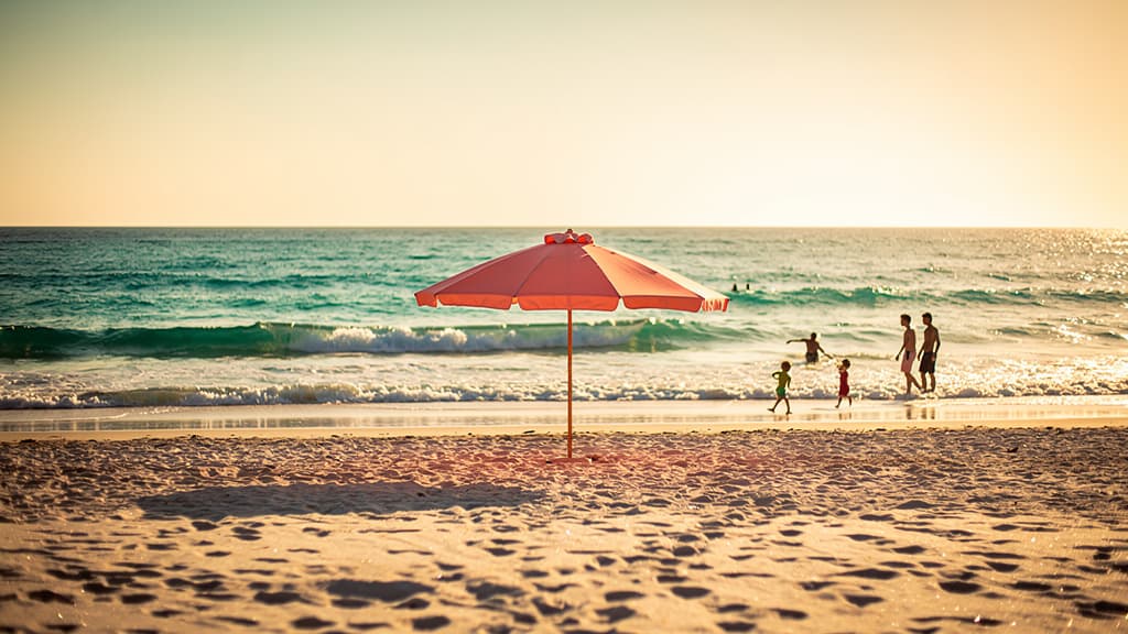 Plage de sable à l’heure dorée, parasol corail et famille au loin près d’une mer turquoise